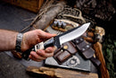 A hand holds the Texan Knives Custom Handmade Skinning Blade with a 4.5" J2 steel blade and buffalo horn & camel bone handle; outdoor gear, ammo, and a leather sheath lie in the background.