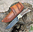 The Texan Knives Custom Handmade 8-Inch Western Knife features a patterned Damascus steel blade and carved handle, shown beside its brown leather sheath outdoors. Crafted by Texan Knives for durability and style.