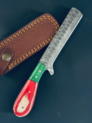 The Texan Knives Bull Cutter Handmade Damascus Steel Knife features a textured, hammered blade and a pakkawood handle with red, green, and white patterns. It comes with a brown leather sheath accented by yellow stitching and a decorative button.