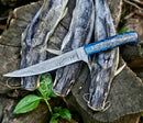 A Texan Knives Custom Handmade Damascus Fillet Knife – 13” with a pine cone handle and leather sheath rests atop three catfish on a rustic wooden surface, accented by green leaves.