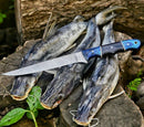 Three freshly caught fish rest on a wooden surface outdoors, with the Texan Knives Custom Handmade Damascus Boning Knife – 13" Pakkawood Handle placed across them, set against a backdrop of green leaves and wooden logs.