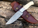 The Texan Knives Custom Handmade Damascus Bushcraft Knife with a patterned blade and camel bone handle rests on its brown leather sheath atop rough wooden logs.
