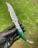 A gloved hand holds the Texan Knives Custom Handmade Damascus Blade EDC Folding Pocket Knife, featuring a 3-inch patterned blade, color bone handle, brass engraved bolster, and leather sheath, set against a blurred grass background.