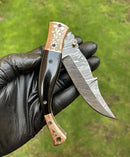 A hand in a black glove holds the Texan Knives Custom Handmade Damascus Blade EDC Folding Pocket Knife featuring a color bone handle and brass engraved bolster, set against a blurred grassy background. Includes leather sheath.
