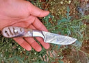 A hand holds the Texan Knives Custom Handmade Damascus Knife in a Belt with a sheep horn handle, set against green plants and tiny white flowers.