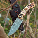 The Texan Knives Custom Handmade Damascus Skinning Knife features a 4.5" patterned blade and a multi-colored sheep horn handle, shown outdoors on tree branches with its brown leather sheath in the background.