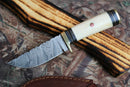 The Texan Knives Custom Handmade Damascus Camping Knife features a patterned blade and a camel bone handle with dark bands and a red inlay, shown on its wooden sheath against a camouflage background.
