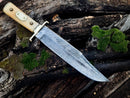 The Texan Knives Damascus Steel Cowboy Bowie Knife – 13" features a brass guard, camel bone handle, and comes with a leather sheath. It rests on dark, weathered wood with green moss patches.