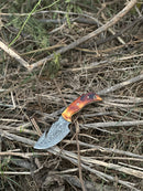 The Texan Knives Custom Handmade Damascus Steel Guthook Skinning Knife features a 7.5” blade, a camel bone handle, and comes with a handcrafted leather sheath, displayed outdoors on dry twigs and grass.