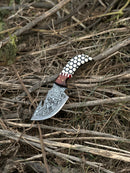 The Texan Knives Custom Handmade Damascus Steel Guthook Skinning Knife (7.5”), featuring a rosewood & resin handle, rests on dry twigs and brown grass next to its leather sheath.