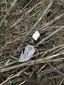 A Texan Knives Custom Handmade Damascus Steel Guthook Skinning Knife, 7.5” with a rosewood, camel bone & black sheet handle and leather sheath, lies among dry grass and twigs outdoors.