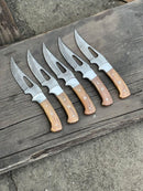Five Custom Handmade Damascus Steel Gutting Knives by Texan Knives, each with a patterned blade and Rosewood handle, are arranged on a weathered wooden surface with a concrete floor in the background.