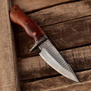 The Texan Knives Custom Handmade Small Hunting Knife features a Damascus steel blade and polished wooden handle, displayed diagonally on a rustic wooden surface.