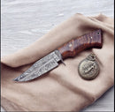The Texan Knives Texas Knives Custom Handmade Damascus Steel Hunting Knife with a rosewood handle and leather sheath is shown on beige fabric beside an ornate silver medallion, all set against a light wood surface.