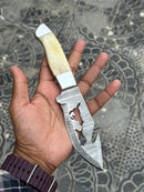 A hand displays the Custom Handmade Damascus Steel Knife in a Belt by Texan Knives, featuring a patterned blade, gut hook, decorative cutout, and camel bone handle against a textured stone background.