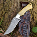 The Texan Knives Premium Damascus Steel Skinner Knife, custom handmade with a camel bone handle, rests on its brown leather sheath beside a tree trunk and green grass.