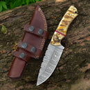 A Texan Knives Custom Handmade Damascus Steel Skinner Knife with a 4.5" patterned blade and polished sheep horn handle rests on a tree trunk next to its brown leather sheath with black snaps and decorative stitching.