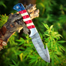 The Texan Knives Custom Handmade Damascus Skinning Knife, featuring an American flag handle, is shown outdoors on a tree branch surrounded by lush green foliage.