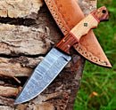 The Texan Knives Custom Handmade Damascus Skinning Knife with a 4.5" patterned steel blade and an olive and rosewood handle rests on rough tree bark beside its tan leather sheath, with grass visible in the background.