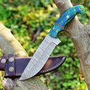 The Custom Handmade Damascus Skinning Knife – 4.5" Resin Handle by Texan Knives rests on a brown leather sheath atop tree branches outdoors, showcasing its green and blue handle perfect for seasonal hunts.