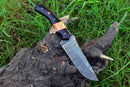 The Texan Knives Custom Handmade Skinning Knife, featuring a Damascus steel blade with distinctive wavy patterns and a handle crafted from buffalo horn and olive wood, rests on a weathered tree stump surrounded by green grass.