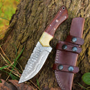 The Texan Knives Custom Handmade Skinning Knife with a Damascus steel blade rests on textured tree bark, featuring a cream and wood handle and leather sheath, surrounded by green grass.