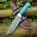 The Texan Knives Custom Handmade Damascus Skinning Knife – 4.5" Blade features a blue patterned handle and rests on a brown leather sheath atop tree logs, surrounded by green leaves.