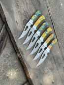 Six Texan Knives Custom Handmade Damascus Skinning Knives with vibrant resin handles are arranged in a row on weathered wood, blades left. The rough concrete background highlights each Texan Knives blade’s artisanal craftsmanship.