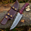 The Texan Knives Custom Handmade 4.5-Inch Damascus Steel Skinning Knife with a patterned blade and durable G10 handle rests on a log next to its brown leather sheath, featuring decorative stitching and brass buttons.