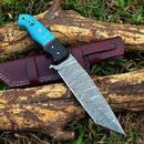 The Texan Knives Custom Handmade Damascus Steel Skinning Knife features a 4.5" patterned blade and a blue resin handle, displayed on its brown leather sheath atop tree branches and green grass.
