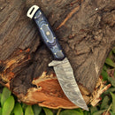The Texan Knives Custom Handmade Damascus Steel Skinning Knife with a 5" blade features a textured handle and wavy pattern, displayed on a rugged tree stump surrounded by green leaves.