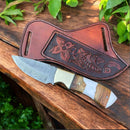 The Texan Knives Custom Handmade Damascus Steel Skinning Knife 7.75", featuring a patterned blade and wood-and-white handle, rests on an embossed leather sheath by Texan Knives, displayed on wood beside green leaves.