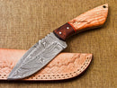 The Texan Knives Custom Handmade Damascus Steel Survival Knife features a 5" patterned blade and a polished wooden handle, resting on a tan leather sheath with decorative stitching and embossed designs.