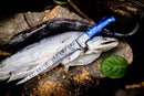 A long, silver fish rests on a tree stump with the Texan Knives Custom Handmade Angling Knife – Damascus Steel Blade & Resin Handle nearby. A second, darker fish and scattered fishing gear surround this premium 2025 fishing tool.