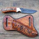 The Texan Knives Buckaroo Belt Knife, featuring an 8" handmade rasp steel blade and an orange epoxy pine cone handle, rests above a tooled Western leather sheath with floral and fish designs on a wooden surface.