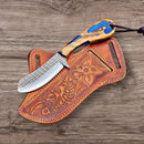 The Texan Knives Custom Handmade Belt Knife features a rasp tool blade and olive wood handle, resting on a tooled leather sheath with floral and fish designs—making it a unique everyday carry item displayed on wood.