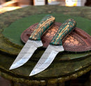 Two Custom Handmade Belt Knives by Texan Knives, each with a 9" patterned rasp tool steel blade and green, gold-accented handles, rest on a brown leather sheath atop a decorative green wooden surface.