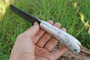 A hand holds the Texan Knives Custom Handmade Cowboy Bull Cutter Knife, showcasing its 4" textured dark J2 steel blade and smooth white handle with black geometric patterns against a backdrop of green grass and wood.