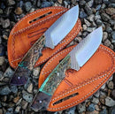 Two Cowboy Bull Cutter Knives by Texan Knives feature custom handmade J2 steel blades and vibrant textured handles, resting on brown leather sheaths atop small rocks.