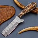 The Texan Knives Cowboy Bull Cutter Knife features a handmade Damascus steel blade, a textured Pakka wood handle, and comes with a brown tooled leather sheath, shown resting on a dark surface near curved antlers.