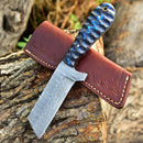 The Texan Knives Cowboy Bull Cutter features a 4" handmade Damascus steel blade and a textured blue-black handle, resting on a brown leather sheath atop tree bark—an eye-catching outdoor piece.