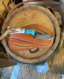 The Texan Knives Custom Handmade Cowboy Knife features a J2 steel blade and a stag horn handle, displayed in a belt sheath on a wooden surface.