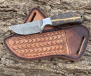 The Texan Knives Winter Edition – a custom handmade Cowboy Knife by Texan Knives, featuring a patterned Damascus steel blade and multicolored handle, rests on its belt rope leather sheath atop textured tree bark.