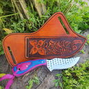 A Texan Knives Custom Handmade Cowboy Knife, 8" with a pink-and-blue Pakkawood handle, rests beside a brown leather sheath embossed with flower and fish designs, surrounded by green plants.