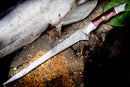 The Texan Knives Handmade Damascus Steel Fish Fillet Knife – 13” with a rosewood handle and leather sheath lies on a wooden surface beside a large, cleaned fish and wood shavings.