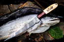 A large fish rests on wood with the Texan Knives Custom Handmade Filleting Knife for Fish—featuring a 13" Damascus steel blade and engraved camel bone & rosewood handle—placed on top. A green leaf sits nearby.