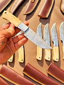 A hand holds the Texan Knives Custom Handmade Damascus Skinner Knife with a camel bone handle and textured blade. Similar Texan Knives and brown leather sheaths sit on a brown surface—an ideal premium hunting gift.