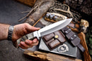 A person with a watch holds the Texan Knives Custom Handmade J2 Steel Skinner Knife with a sheep horn handle. Texan Knives outdoor gear, wallet, ammo, and survival items are arranged on a wooden surface in the background.