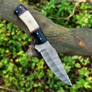 A Texan Knives Custom Handmade Skinning Knife with a 4.5" Damascus blade and a camel bone & resin handle rests on a tree branch, with green foliage blurred in the background.