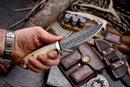 A hand holds the Texan Knives Custom Handmade Skinning Knife with a 4.5" patterned Damascus steel blade and camel bone handle, surrounded by shotgun shells, a leather pouch, gun holster, and hunting gear.
